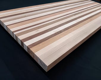 May include: A rectangular wooden cutting board featuring a striped pattern composed of alternating light, medium, and dark brown wood planks. The surface displays a smooth, flat finish with visible natural wood grain textures running lengthwise.