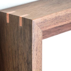 May include: A dark brown wooden picture frame featuring a mitered corner joint reinforced with two light-colored rectangular splines. The frame has a visible wood grain texture and a smooth finish, showcasing the natural dark tones of the wood.