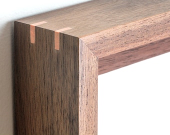 May include: A dark brown wooden picture frame featuring a mitered corner joint reinforced with two light-colored rectangular splines. The frame has a visible wood grain texture and a smooth finish, showcasing the natural dark tones of the wood.
