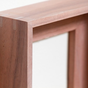 May include: A rectangular wooden picture frame with a natural reddish-brown wood grain finish. The frame features mitered corners with visible joinery details, showcasing a deep profile and a smooth, matte surface texture throughout the structure.