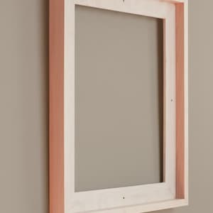 May include: A rectangular wooden picture frame with a light natural wood finish on the front face and a contrasting reddish-brown wood tone on the side edges. The frame features visible finger joint construction at the corners and small pre-drilled holes.