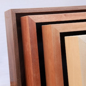 May include: Close-up of a stack of wooden picture frames in various shades of brown and beige. The frames are stacked, showing the corners and edges, with a dark brown frame on top, followed by reddish-brown, light brown, and a cream-colored frame.