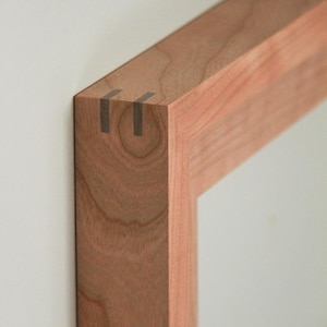 May include: Close-up of a wooden frame corner. The frame is constructed from light brown wood, with visible grain and a smooth finish. The corner features a dovetail joint, with dark gray inlays. The frame is mounted on a white wall.