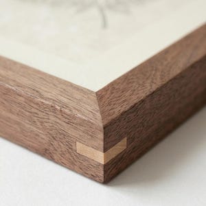 May include: Close-up of a wooden picture frame with a light-colored drawing inside. The frame is made of dark brown wood with a light-colored wood accent at the corner joints. The frame is square and has a clean, modern design.