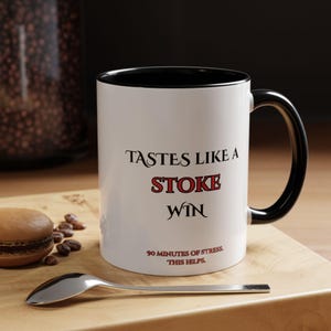 Puede incluir: Taza de cerámica blanca con interior y asa negros. La taza presenta el texto "TASTES LIKE A STOKE WIN" en negro y rojo, con "90 MINUTES OF STRESS, THIS HELPS" debajo. Una cuchara y un macaron están sobre una superficie de madera.