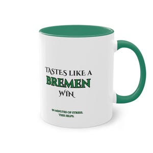 Puede incluir: Taza de cerámica blanca con interior y asa verdes. La taza presenta el texto "TASTES LIKE A BREMEN WIN" en negro y verde, con el texto adicional "90 MINUTES OF STRESS. THIS HELPS."