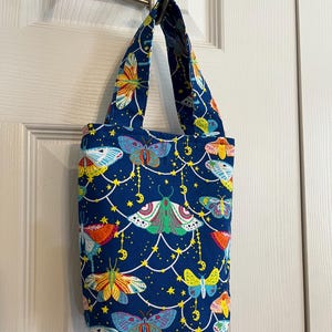 May include: A small blue tote bag with a colorful butterfly and moon pattern. The bag has two straps and is hanging on a hook.