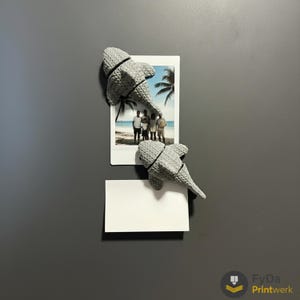 Può includere: Due calamite grigie a forma di squalo sono attaccate a una superficie grigia, che tengono una foto e una scheda bianca vuota. Le calamite hanno un aspetto strutturato e sono posizionate sopra e sotto la foto. La foto mostra una famiglia sulla spiaggia.