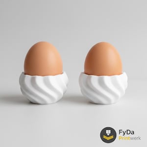 Peut inclure: Deux œufs bruns reposent dans des coquetiers blancs et tourbillonnants. Les coquetiers ont un design moderne et minimaliste. Le fond est gris clair uni. Le logo FyDa Printwerk est en bas à droite.