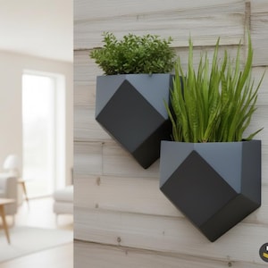 Op de afbeelding: Twee geometrische, aan de muur gemonteerde plantenbakken in donker- en lichtgrijs, elk met groene planten. Ze zijn bevestigd aan een lichtgekleurde houten muur. Het Fyda Werk-logo is rechtsonder zichtbaar.