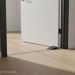 Può includere: Un fermaporta a cuneo nero, lungo 12,7 cm, alto 3 cm e largo 15,2 cm, è posizionato su un pavimento in legno chiaro, tenendo socchiusa una porta bianca. Il telaio della porta è nero. Il testo "FyDa Printwerk" è visibile.