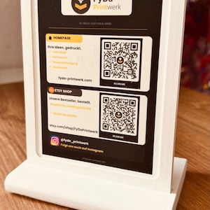 Könnte beinhalten: Ein weißer, 3D-gedruckter rechteckiger Schilderständer mit einer schwarzen Infokarte. Die Karte zeigt das FyDa Printwerk Logo, deutschen Text und zwei QR-Codes mit der Aufschrift 'SCAN ME' für eine Homepage und einen Etsy-Shop sowie einen Instagram-Namen.