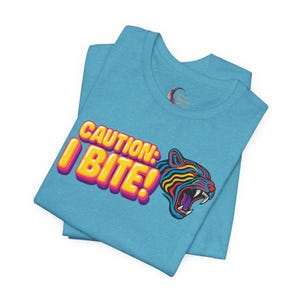 Puede incluir: Una camiseta azul claro con el texto "CAUTION: I BITE!" en una fuente retro colorida. La camiseta presenta un gráfico de un tigre gruñendo con rayas vibrantes. La camiseta está doblada.