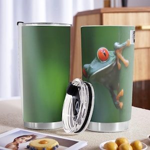 May include: Two stainless steel travel mugs. One is solid green, the other features a close-up of a red-eyed tree frog on a green background. A clear lid with a black seal sits in front of the mugs. The mugs are on a light-colored surface.