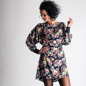May include: A floral print dress with a dark navy blue background. The dress features a boat neckline, long sleeves with navy cuffs, and a fitted waist. The floral pattern includes pink, yellow, and white flowers.