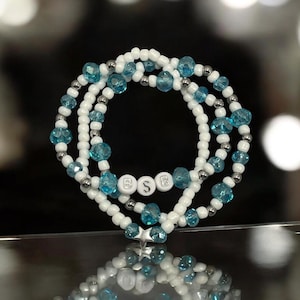 Op de afbeelding: Een stapel van drie armbanden met kralen. De armbanden hebben witte en zilveren kralen, afgewisseld met heldere blauwe facetkralen. De middelste armband heeft de letters "B S B" in witte letterkralen. De armbanden zijn op elkaar gestapeld.