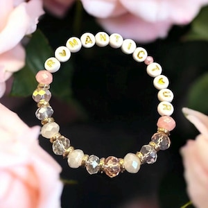 Op de afbeelding: Een armband met het woord "ROMANTIC" gespeld in gouden letters op witte kralen. De armband heeft roze en heldere kristallen kralen, met gouden accenten. De achtergrond is een wazige afbeelding van roze rozen.