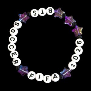 Könnte beinhalten: Ein kreisförmiges Perlenarmband mit weißen runden Perlen, die mit schwarzen Buchstaben die Wörter BTS, SOCCER, FIFA und 2026 bilden. Das Armband ist mit durchscheinenden, glitzernden violetten sternförmigen Perlen und kleinen schwarzen Zwischenperlen versehen.