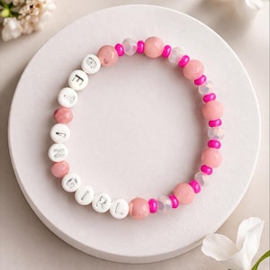 May include: A pink and white beaded bracelet with the word "GIRL" spelled out in white letter beads. The bracelet features pink and clear beads, with a white background and floral accents.