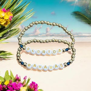 Puede incluir: Dos pulseras de cuentas doradas con cuentas de letras blancas que deletrean "FLORIDA" y "BAHAMAS". Las pulseras se exhiben sobre una superficie arenosa con follaje tropical y una escena de playa de fondo. Las pulseras tienen detalles azules.