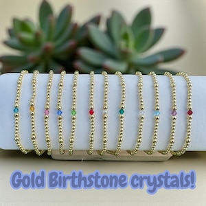 May include: A collection of gold beaded bracelets, each featuring a different colored birthstone crystal. The bracelets are displayed on a white surface with a succulent plant in the background. The text "Gold Birthstone crystals!" is at the bottom.