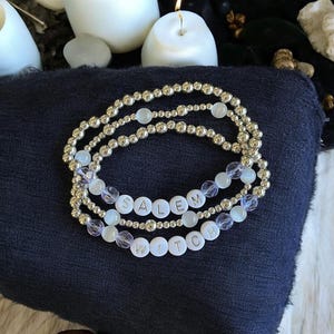 May include: Three beaded bracelets with the words "SALEM WITCH" spelled out in white letter beads. The bracelets are silver and feature clear and light blue beads. The bracelets are displayed on a dark blue surface.