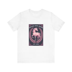 Könnte beinhalten: Weißes T-Shirt mit einer rosa Pferdeillustration in einem dunkelblauen Rechteck, umgeben von einem Lorbeerkranz. Der Text "BRUSH HER MANE" steht über dem Pferd, darunter "EST. 2006".