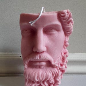 May include: A pink candle in the form of a classical male bust, featuring a beard and curly hair. The candle has a white wick and is set against a plain backdrop. The bust is a detailed sculpture.
