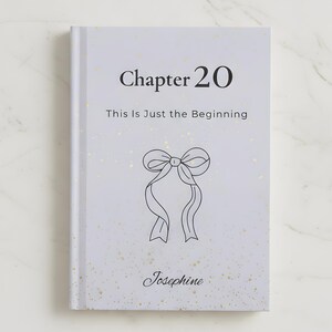 May include: A light gray journal with the text "Chapter 20 This Is Just the Beginning" and a bow illustration. The name "Josephine" is written below the bow. The cover has a subtle gold speckled design.
