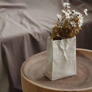 May include: A white ceramic vase with a paper bag design holds white daisy-like flowers and brown leaves. The vase sits on a round wooden tray, with a neutral-toned fabric in the background.