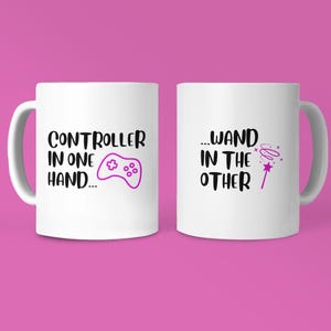 May include: Two white ceramic mugs with black text and purple graphics. One mug reads "CONTROLLER IN ONE HAND..." with a game controller graphic. The other mug reads "...WAND IN THE OTHER" with a magic wand graphic. The background is pink.