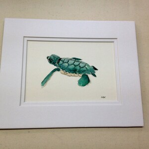May include: A framed watercolor painting of a small, green sea turtle swimming. The turtle is painted in shades of green and blue, with a white underbelly. The painting is signed "LAK" in the bottom right corner.