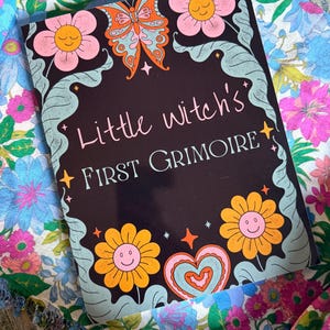 May include: A black book cover with the text "Little Witch's First Grimoire" in pink script. The cover features a butterfly, flowers, and a heart in orange, pink, and blue. The book is set against a floral patterned background.