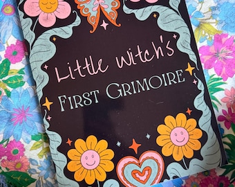 Puede incluir: Una portada de libro negra con el texto "Little Witch's First Grimoire" en escritura rosa. La portada presenta una mariposa, flores y un corazón en naranja, rosa y azul. El libro está sobre un fondo con estampado floral.