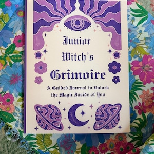 May include: A purple and white "Junior Witch's Grimoire" journal with celestial illustrations, including planets and a crescent moon. The cover features an eye design and floral accents. The journal is on a floral patterned fabric.