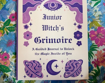 Puede incluir: Un diario morado y blanco "Junior Witch's Grimoire" con ilustraciones celestiales, incluyendo planetas y una luna creciente. La portada presenta un diseño de ojo y detalles florales. El diario está sobre una tela estampada floral.