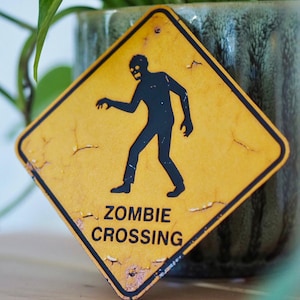 Könnte beinhalten: Ein gelbes, rautenförmiges Schild mit schwarzem Rand und abgenutztem Finish. Das Schild zeigt eine schwarze Silhouette eines Zombies und die Worte "ZOMBIE CROSSING" in schwarzer Schrift. Das Schild lehnt an einem grün-grauen Keramiktopf.