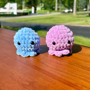 May include: Two small, round, crocheted plush toys shaped like octopuses. One is light blue and the other is light purple. Both have textured, yarn-based surfaces, small black plastic eyes, and multiple short, rounded tentacles at the base.