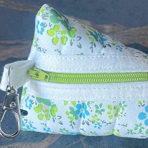 May include: A triangular-shaped, floral-patterned pouch with a lime green zipper and a silver-colored clasp. The pouch features a white background with a repeating pattern of green and blue flowers. The zipper is open, revealing the interior. The pouch is made of fabric.