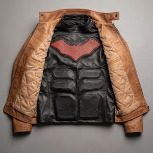 May include: A tan leather jacket with a quilted beige interior lining, worn open over a black leather chest piece. The black piece features a red bat-shaped emblem on the upper chest and molded, padded sections across the torso area.