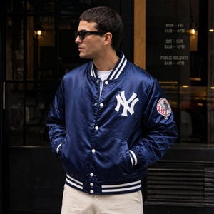 Può includere: Giacca bomber in raso blu navy con strisce bianche su colletto, polsini e orlo. La giacca presenta il logo dei New York Yankees in bianco sul petto e una toppa della squadra sulla manica. La giacca ha una chiusura con bottoni e due tasche.