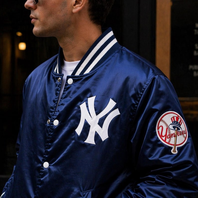 May include: Navy blue satin bomber jacket with white accents and the New York Yankees logo. Features a white "NY" logo on the chest and a Yankees patch on the sleeve. The jacket has a striped collar and white snap closures.
