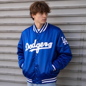 Può includere: Giacca in raso blu reale con dettagli bianchi e la scritta "Dodgers" in bianco sul petto. La giacca ha una chiusura con bottoni a pressione, strisce bianche sui polsini e sull'orlo e le lettere "LA" sulla manica.
