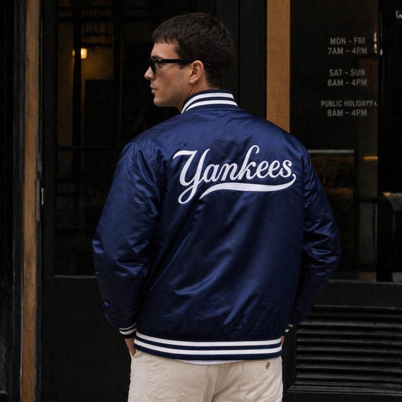 May include: Navy blue satin Yankees baseball jacket with white trim on the collar, cuffs, and hem. The word "Yankees" is written in white script across the back. The jacket is worn by a person wearing sunglasses.