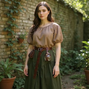 May include: A woman wearing a brown peasant blouse with short, puffed sleeves and a long, olive green skirt. A brown leather belt with a pouch and a glass vial is around the waist. The background features a brick wall and greenery.