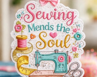 May include: Embroidered sewing-themed wall hanging with the text "Sewing Mends the Soul." Features a teal sewing machine, pink thread spool, yellow measuring tape, and scissors. A pink ribbon and bow are at the top. The design is framed by a white lace edge.
