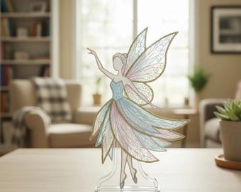 May include: A decorative fairy figurine with a blue, pink, and gold color scheme. The fairy has outstretched arms and detailed wings. The figurine is mounted on a clear base and is displayed on a wooden table.