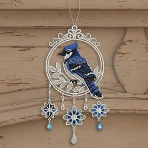 Puede incluir: Adorno decorativo con una arrendajo azul posada en una rama dentro de un círculo de filigrana blanca. Debajo, tres acentos florales azules y blancos con colgantes de lágrima cuelgan. El adorno está diseñado para colgar.