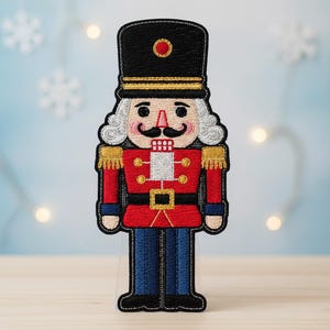 May include: Embroidered patch of a toy soldier nutcracker. The patch features a black hat with a red circle, a red jacket with gold accents, and blue pants. The nutcracker has a white wig and a black mustache.
