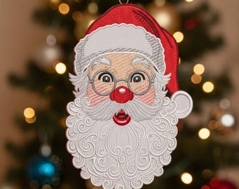 Pode incluir: Enfeite de Natal bordado do Pai Natal, com gorro vermelho, barba branca e bochechas rosadas. O enfeite está pendurado por uma fita vermelha contra um fundo desfocado de uma árvore de Natal decorada.